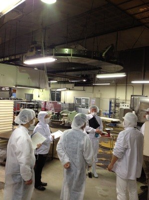 白衣・帽子姿の担当者たちが並ぶ食品製造現場で、担当者がボトルを手に説明している。